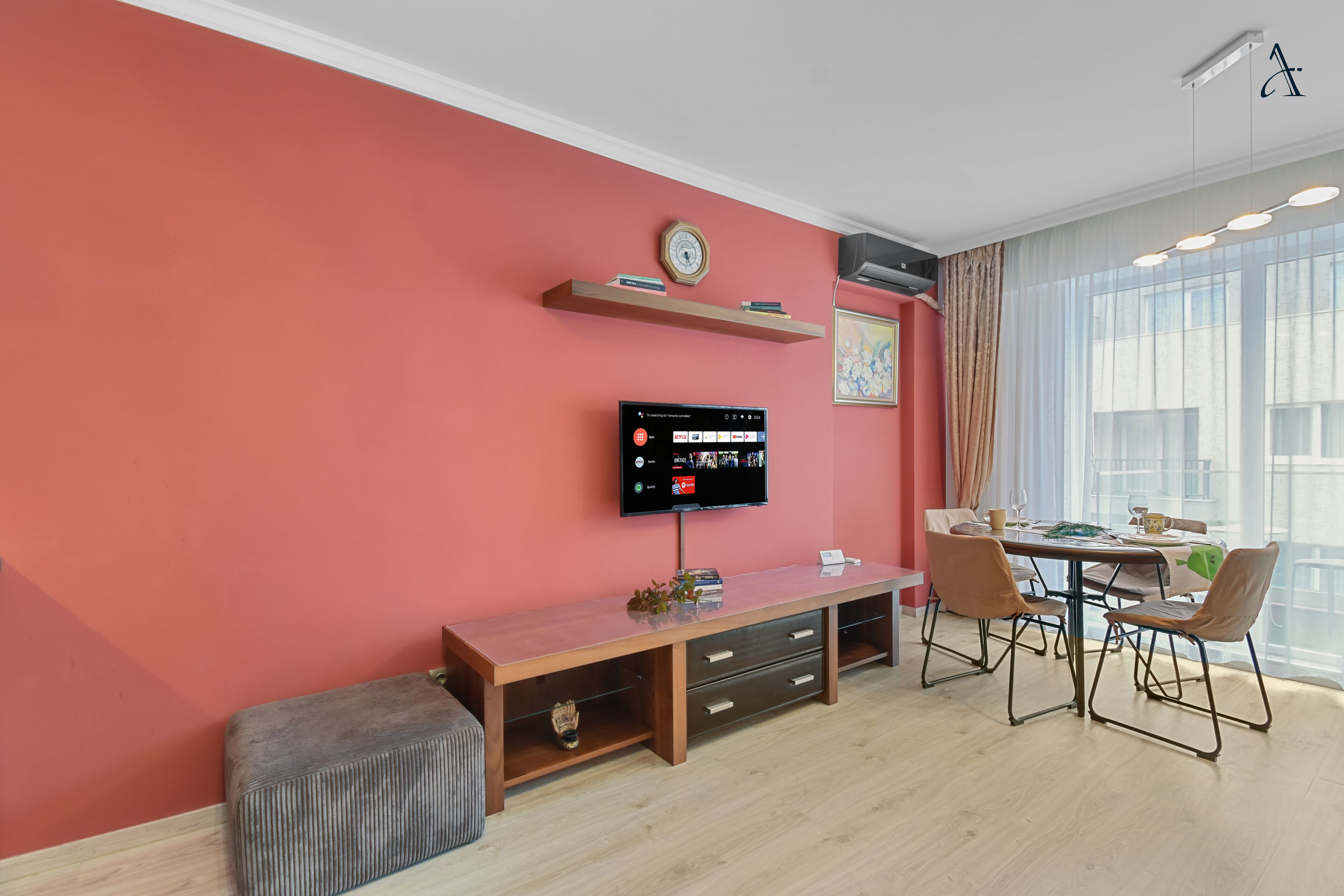 A modern living room with a red accent wall, a flat-screen TV, a wooden bench, a dining table with chairs, and a large window with curtains.