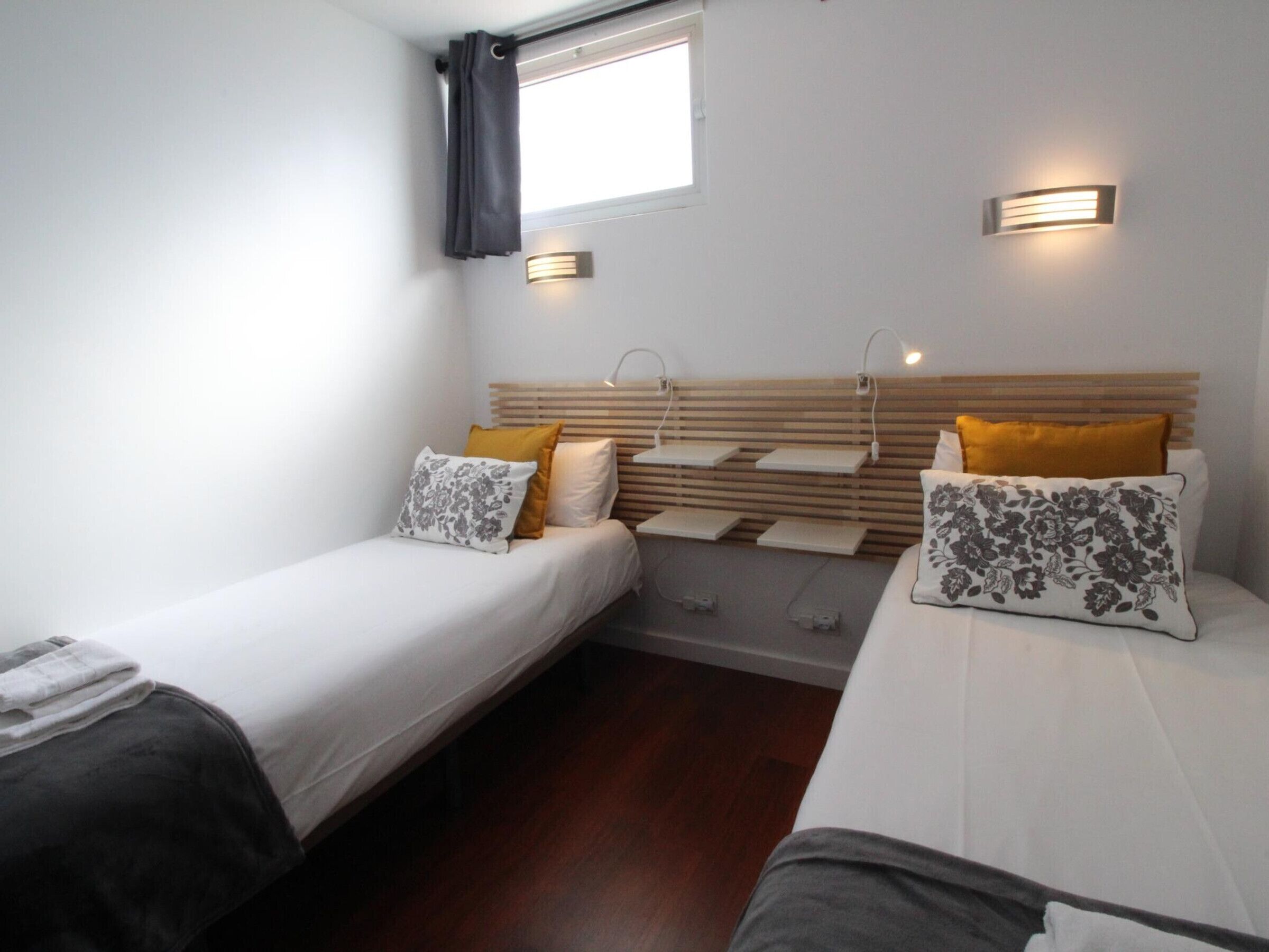 Ein Zimmer mit zwei Betten, jeweils mit einem hölzernen Kopfteil, weißer Bettwäsche und dekorativen Kissen. An den Wänden sind Leuchten angebracht und es gibt ein kleines Fenster mit Vorhang.