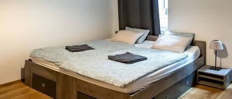 A modern bedroom with a wooden bed frame, a large window with black curtains, and a bedside lamp.