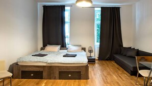 Ein modernes Schlafzimmer mit Holzboden, einem Bett mit grau-weiß gemustertem Überwurf, einem schwarzen Sofa, einem kleinen Tisch und einer Pendelleuchte.