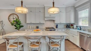 A kitchen with white cabinets, a central island with bar stools, pendant lighting, and a large window with blinds. - Villa De Luxe Near Forsyth Park w/Private Hot Tub (Savannah)