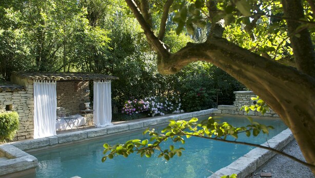 A rectangular pool with stone borders, surrounded by lush greenery and a stone structure with white curtains. - Daudet Romantic two-person apartment in Provence (St Remy de Provence Quartier de Paluds)