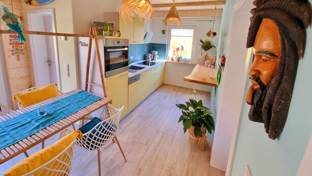 A modern kitchen with wooden cabinets, a built-in oven, and a dining area with a table and chairs. - Ferienwohnung Tiki-beach, Karibisches Flair in der Oberlausitz (Malschwitz)