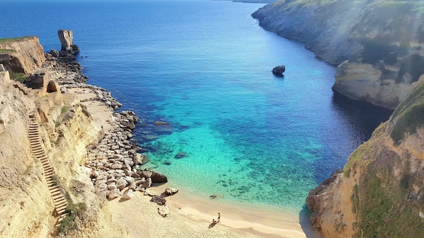 A coastal landscape with a clear blue sea, rocky cliffs, and a sandy beach.