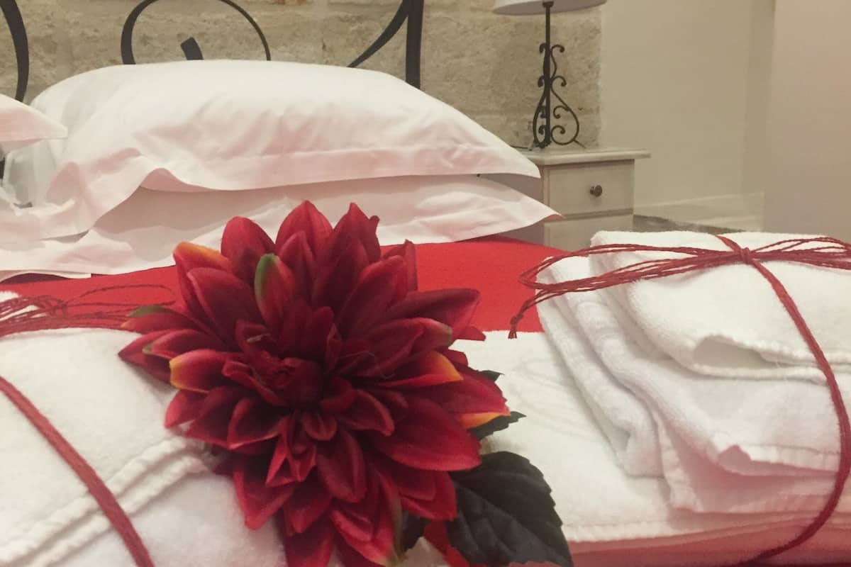 Una cama bien hecha con sábanas blancas y una decoración de flor roja, una mesita de noche blanca con una lámpara, y una pared de ladrillo al fondo.