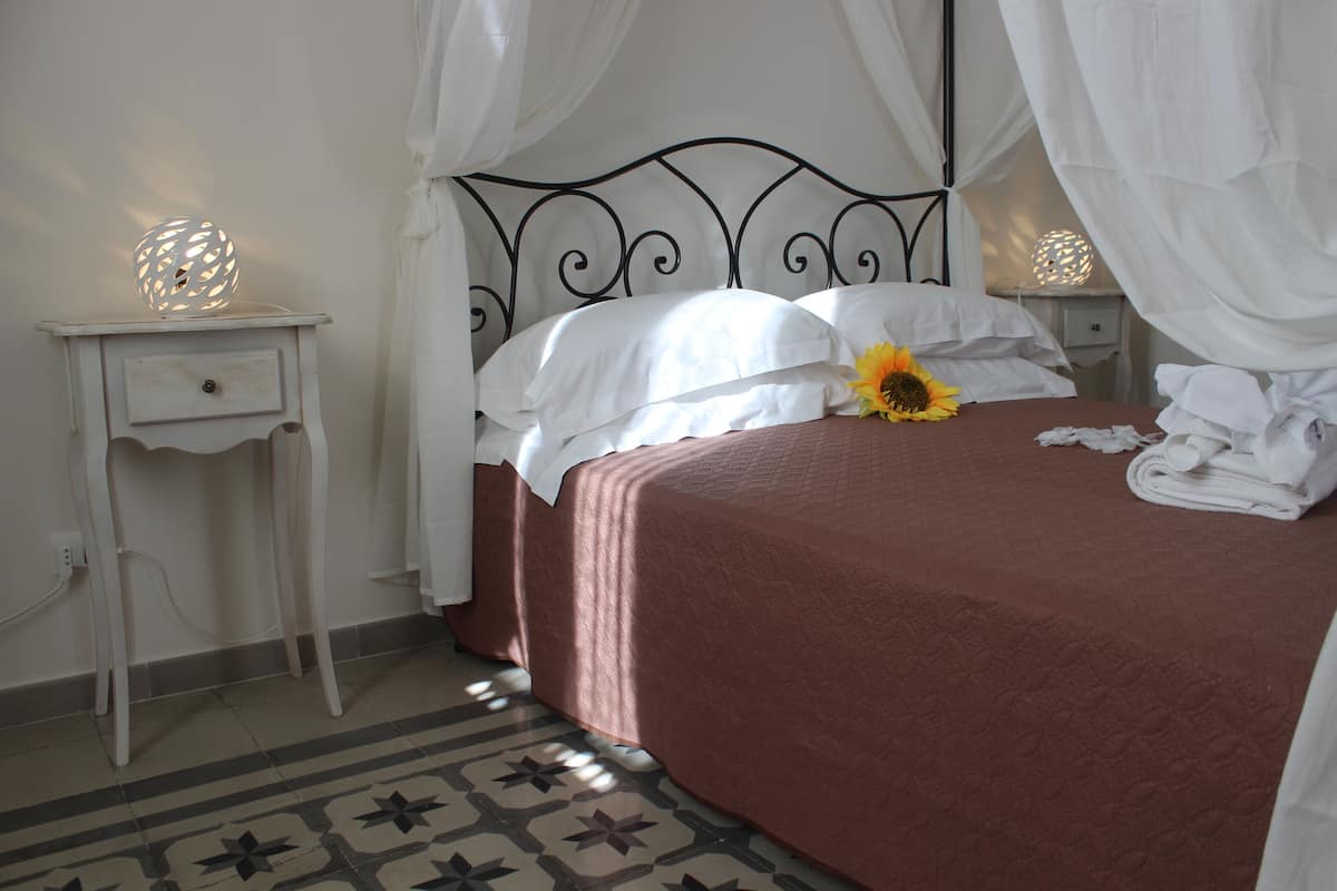 Una cama bien hecha con colcha marrón, almohadas blancas y una sola girasol. Hay dos mesitas de noche con lámparas, una de ellas con luz decorativa. El cuarto tiene un piso estampado y una pared blanca.