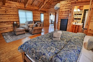A log cabin interior with a large bed, two armchairs, a sofa, a fireplace, and a bathroom with a mirror and shelves.