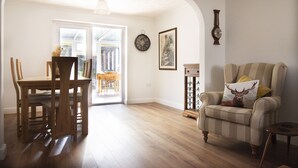 A dining area with a wooden table and chairs, a living room with a striped armchair and a decorative pillow, and a hallway leading to a door. - Bilbo's Cottage, Sheringham (Sheringham)