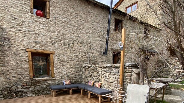Una terraza de madera con un sofá y sillas, un edificio de piedra con ventanas, y un árbol.