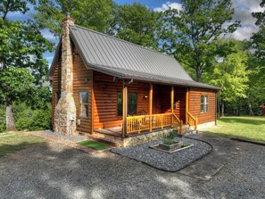 A log cabin with a stone chimney, wooden porch, and a gravel driveway.