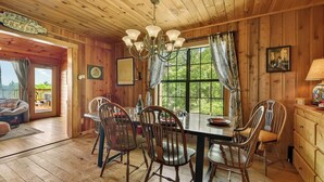 A wooden cabin interior with a dining table, chairs, a chandelier, and a large window with curtains.
