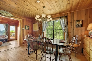 A wooden cabin interior with a dining table, chairs, a chandelier, and a large window with curtains.