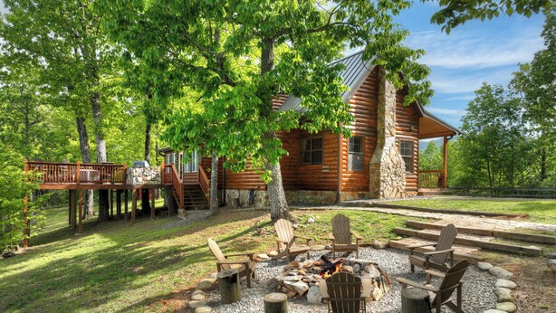 A log cabin with a deck, a fire pit with chairs, and a stone wall.