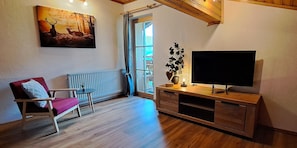 Una sala de estar con un sillón rojo, un mueble de madera con televisión, un cuadro de ciervos y una ventana con cortinas.
