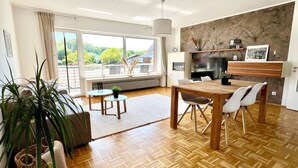 Ein modernes Wohnzimmer mit einem Holzesstisch, weißen Stühlen, einer Couch und einem großen Fenster mit Blick ins Grüne.