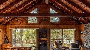 A spacious cabin interior with a high ceiling, wooden beams, a dining area, a sofa, and a stone wall.