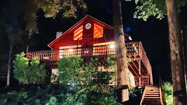 A wooden cabin with a red roof, surrounded by dense greenery and a wooden staircase leading up to it.