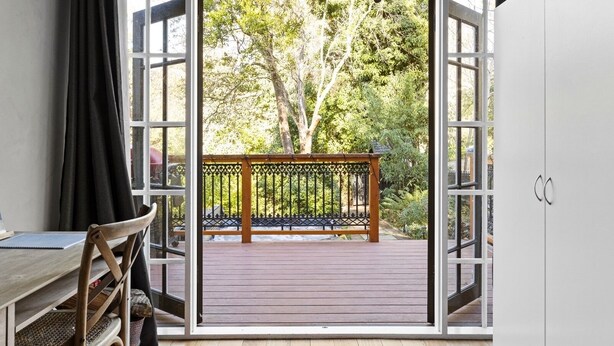 A room with a wooden table and chairs, a bench outside, and a view of a garden through open doors.