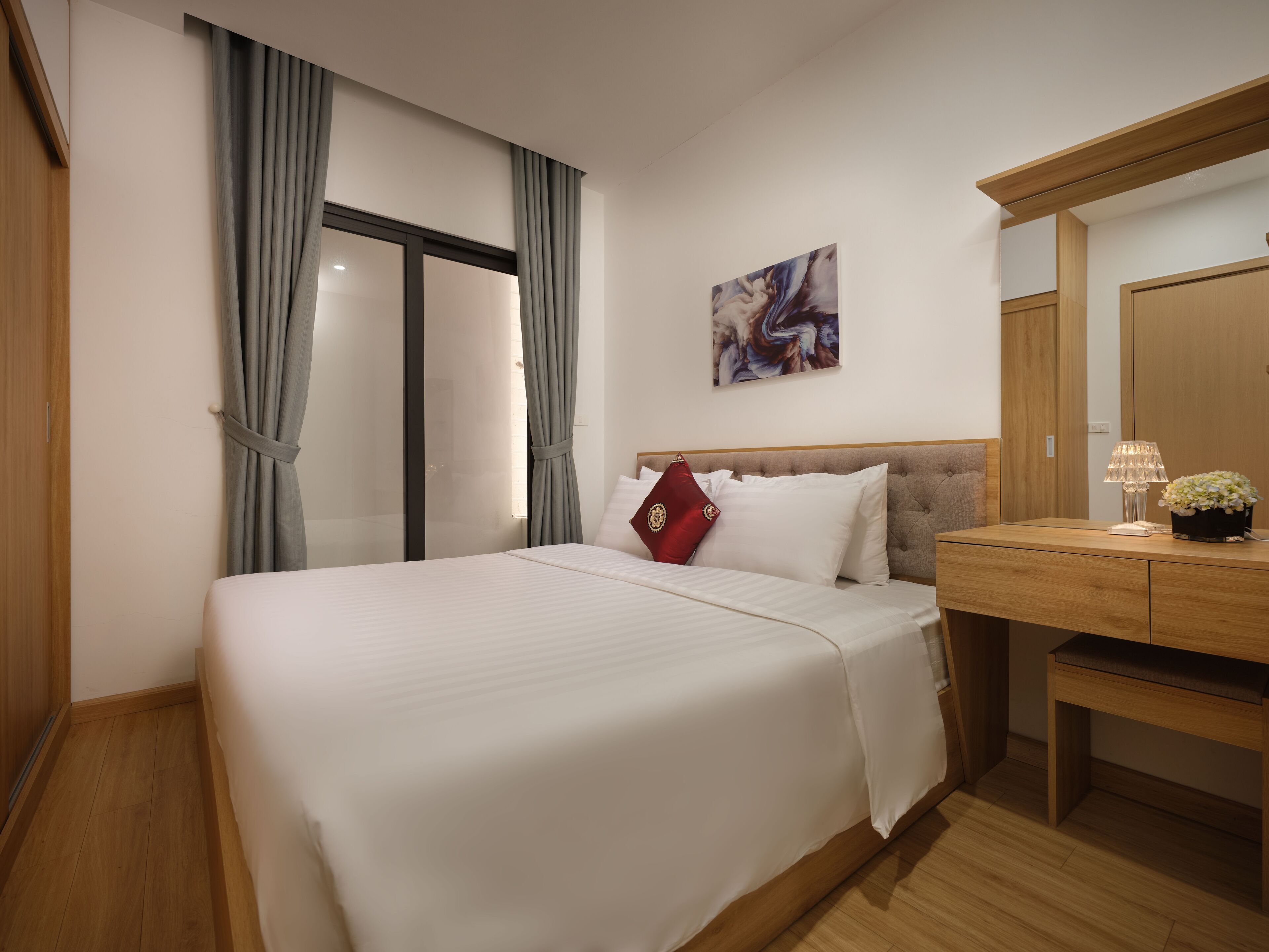 A hotel room with a bed, a wooden desk, a lamp, and a painting on the wall.