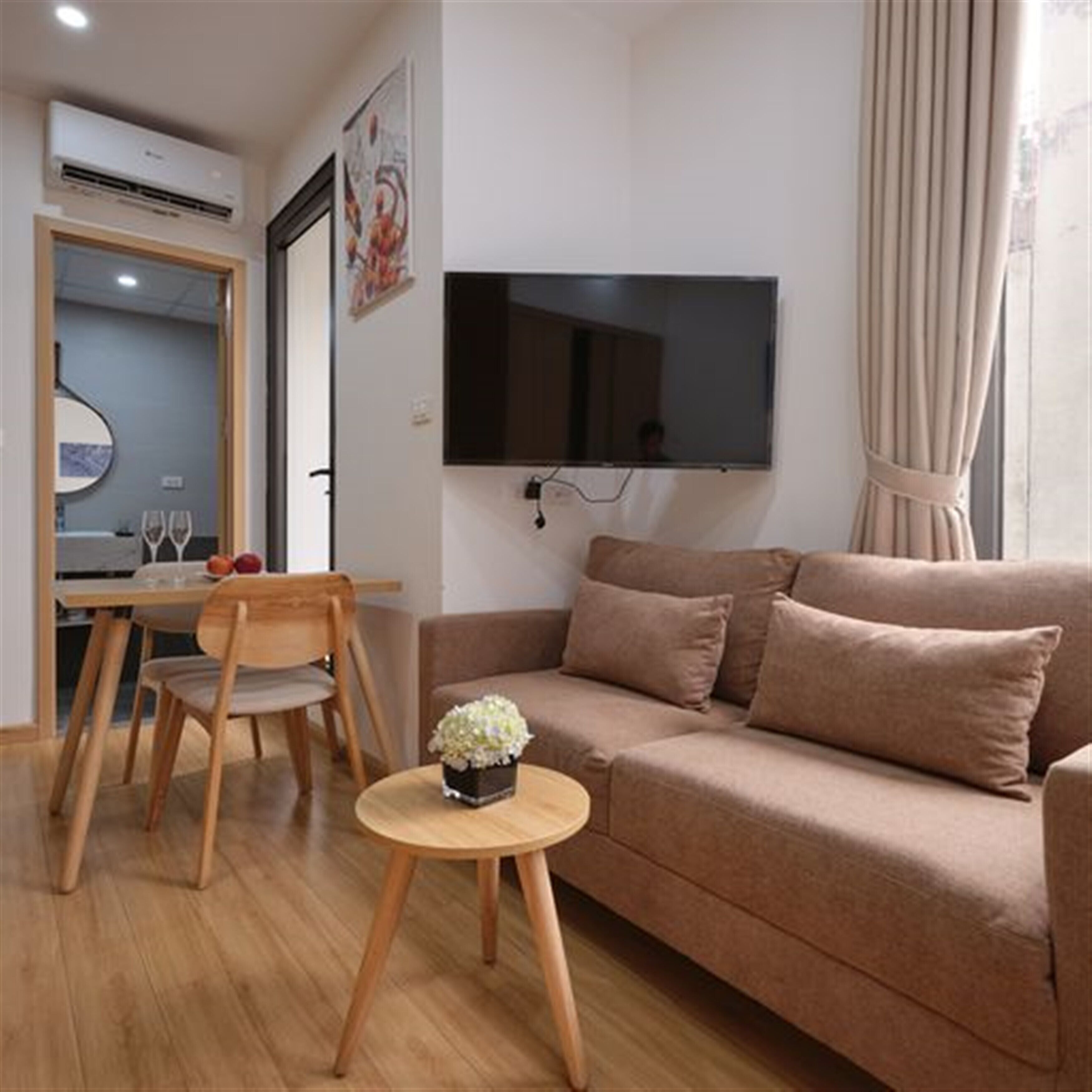Ein Wohnzimmer mit einer Couch, einem an der Wand befestigten Fernseher, einem kleinen runden Tisch mit Blumenvase und einem Essbereich mit Tisch und Stühlen.