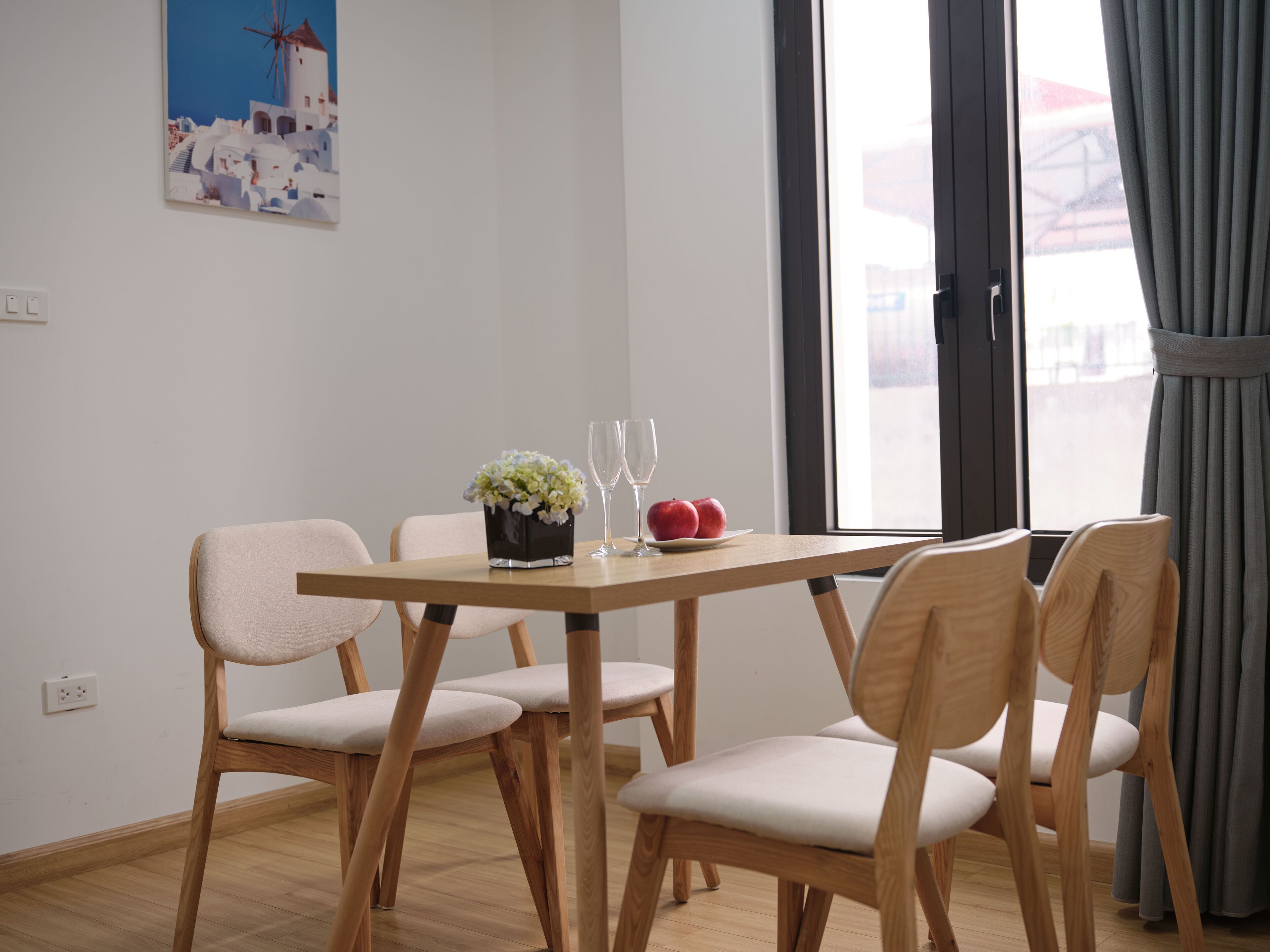 Un comedor con una mesa de madera, cuatro sillas, un jarrón de flores, dos vasos y dos manzanas.