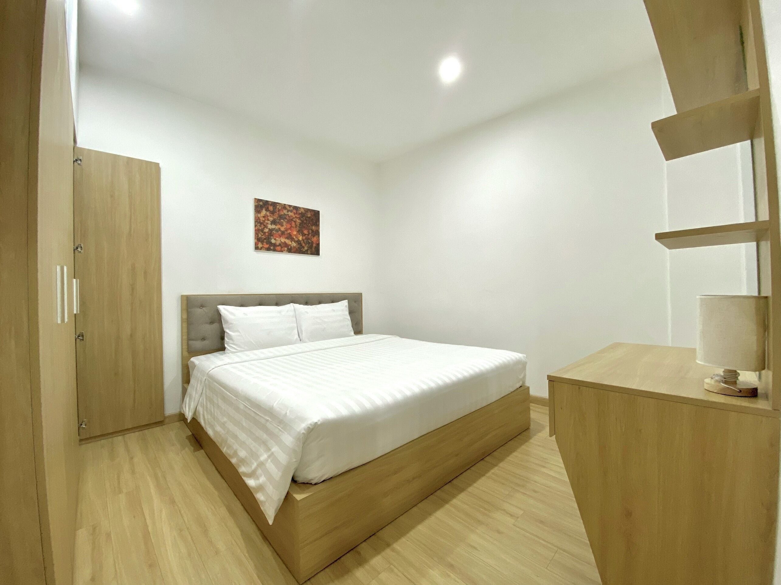 A bedroom with a bed, a wooden wardrobe, a desk with a lamp, and a painting on the wall.