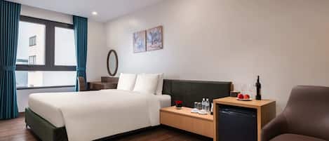 A hotel room with a bed, a wooden nightstand, a small table with a wine bottle, a chair, and a view of buildings through the window.