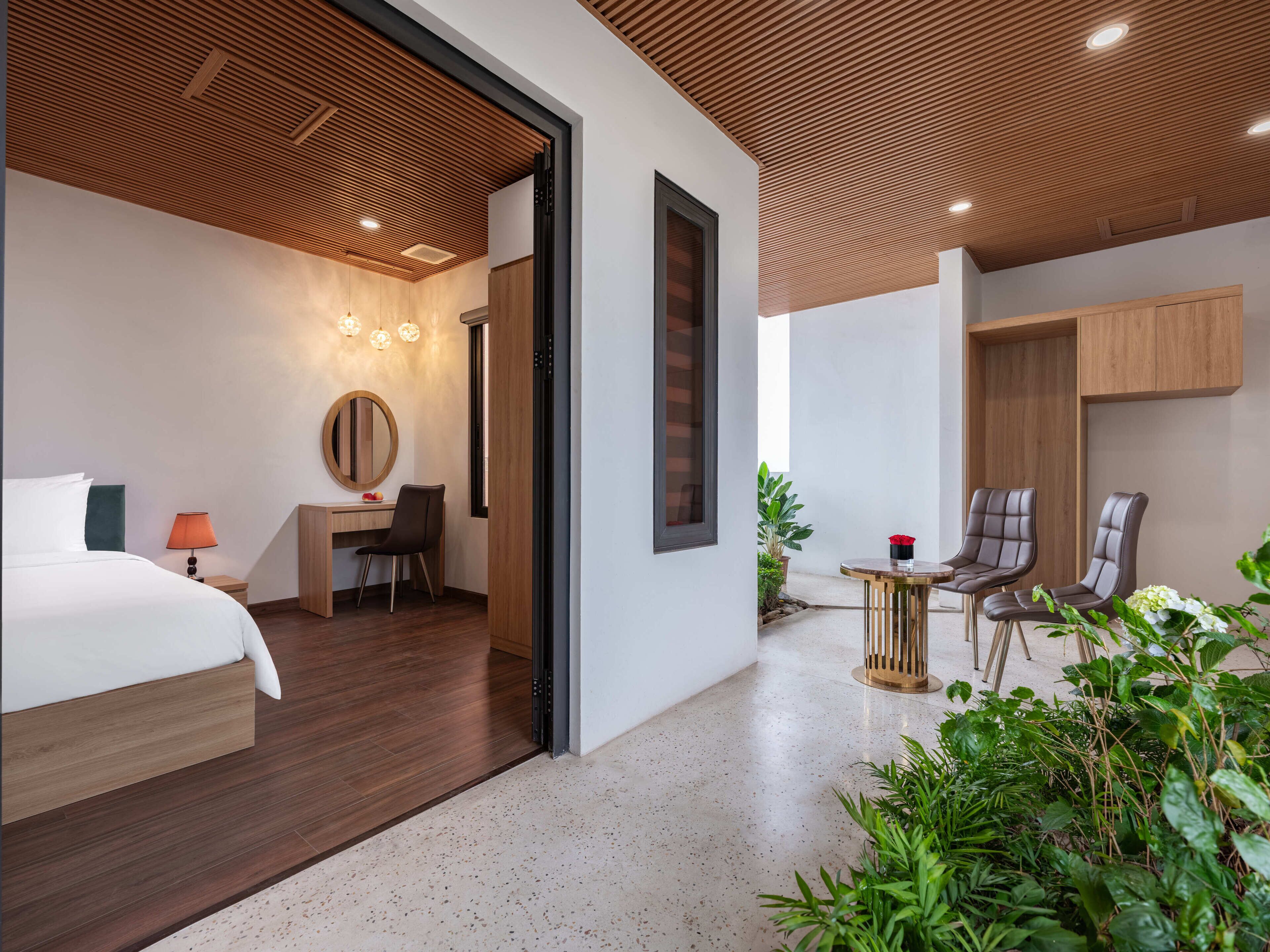 A modern hotel room with a bed, desk, and chairs, a large mirror, and a small balcony with plants.