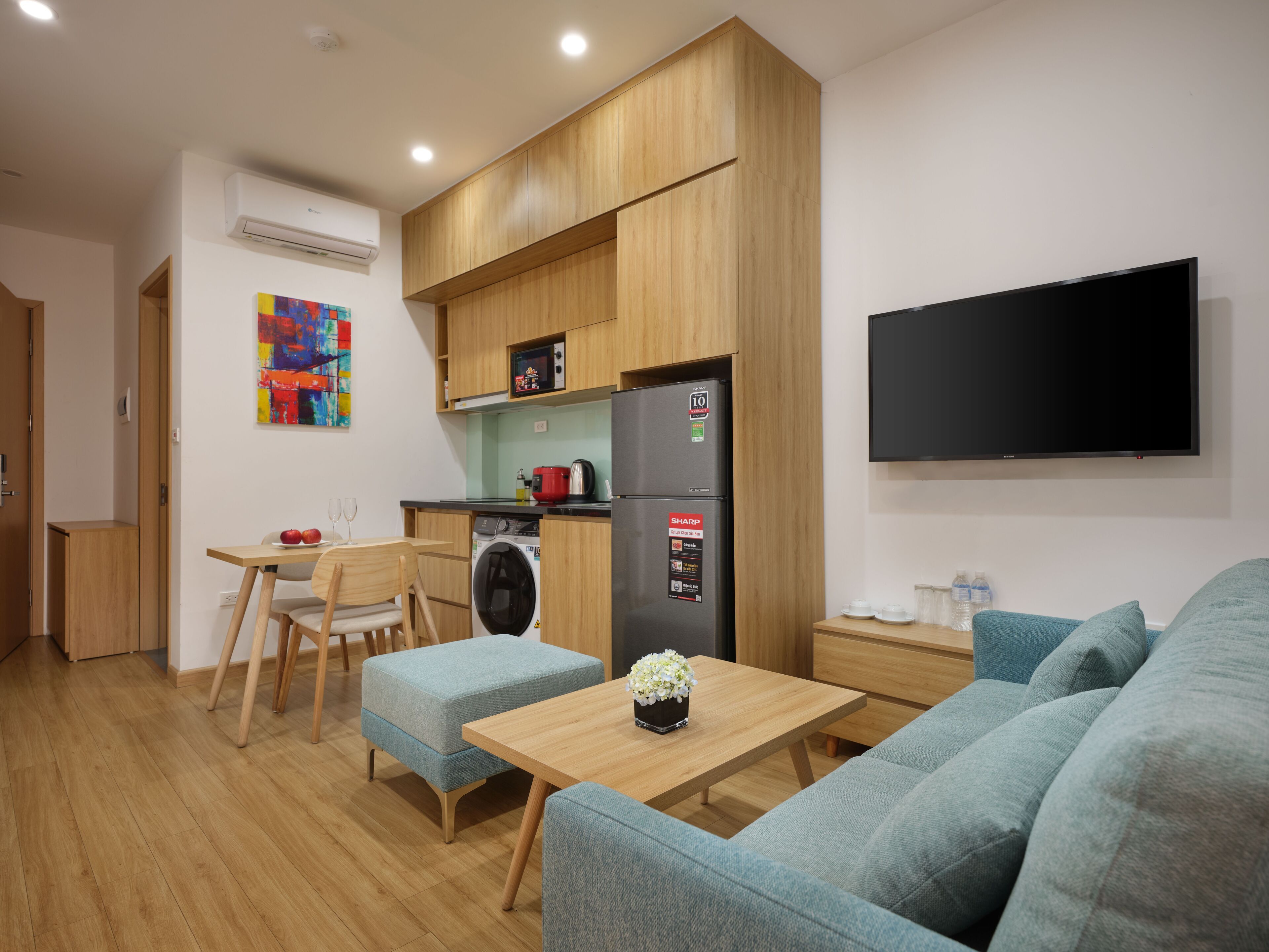 A compact kitchen with wooden cabinets, a washing machine, a refrigerator, and a flat-screen TV.