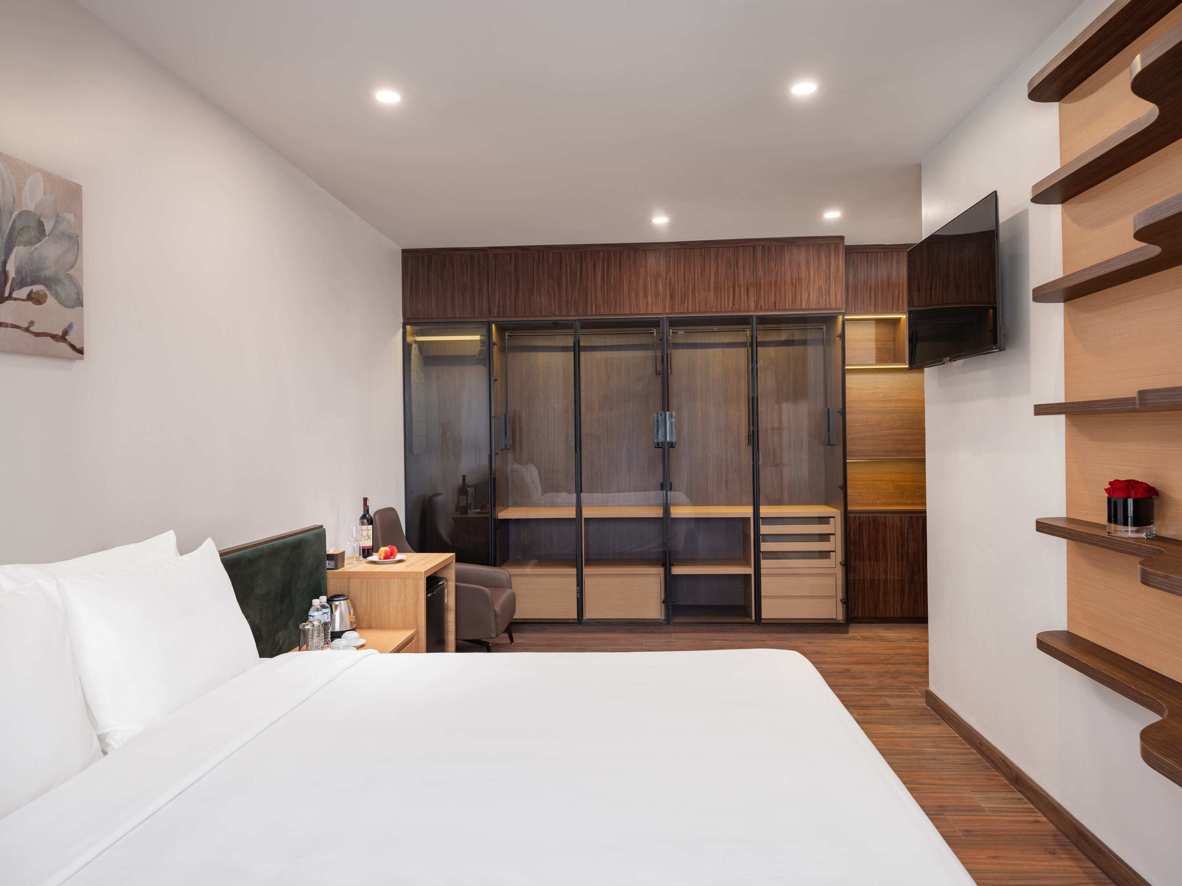 A modern hotel room with a large bed, a desk with a chair, a wardrobe, a television mounted on the wall, and a small table with a bottle and glass.