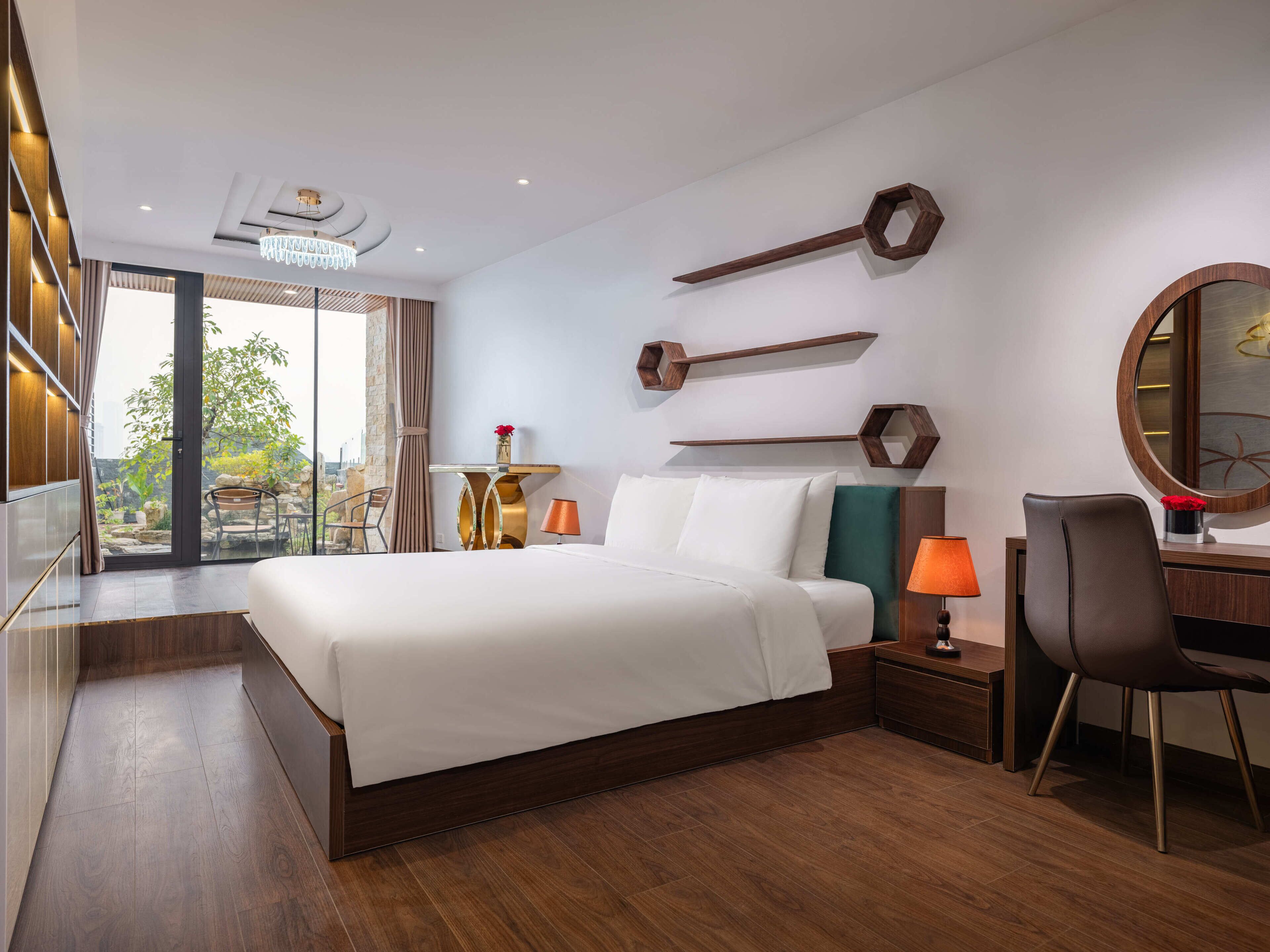 A modern hotel room with a large bed, a desk with a chair, a round mirror, and a view of the outdoors.