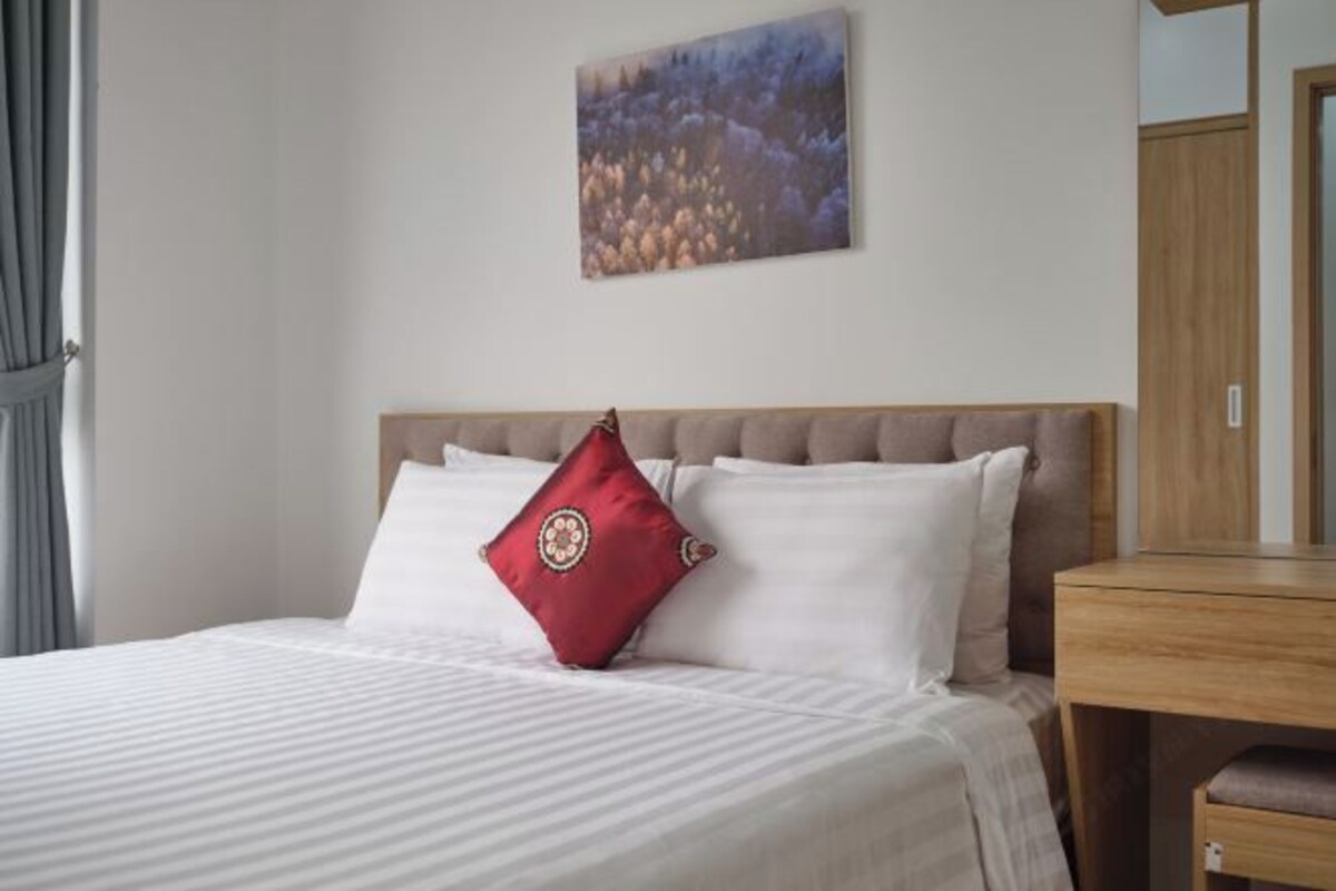 Una cama bien hecha con ropa blanca y una almohada decorativa roja, un cabecero de madera y un cuadro en la pared.
