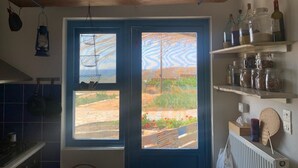 Et køkken med en blå dør, et vindue der viser et landskab, og hylder med glas og køkkenredskaber.