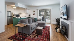 Ein modernes Wohnzimmer mit einem grauen Ecksofa, einem kleinen Holztisch und einem Kamin. Es gibt eine Küchenzeile mit weißen Schränken und einem Edelstahlkühlschrank. Ein großes Fenster bietet einen Blick ins Freie.