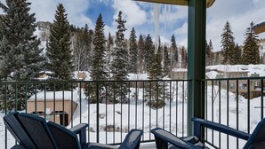Ein Balkon mit zwei blauen Adirondack-Stühlen, einer Holzterrasse und Blick auf einen verschneiten Wald und Gebäude.
