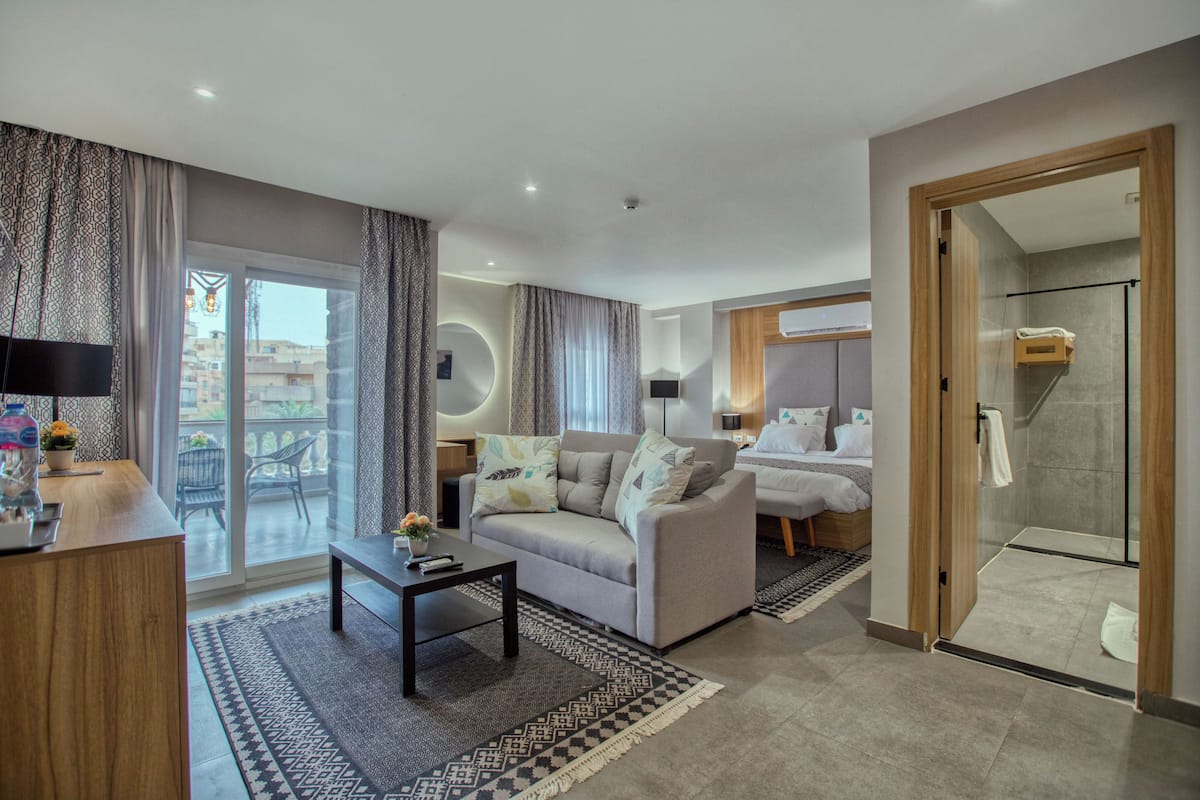 Et moderne hotelværelse med en sofa, et lille bord, en glas brusekabine og en balkon med udsigt til bygninger.
