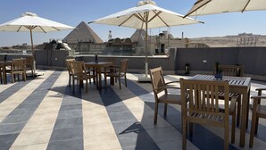 An outdoor seating area with tables and chairs, large umbrellas, and a view of a pyramid structure in the background. - Locanda Museum Hotel (Giza)