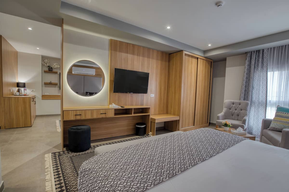 Et moderne hotelværelse med en stor seng, et TV monteret på en trævæg, et skrivebord med en stol og et opholdsområde med en stol og en sofa.