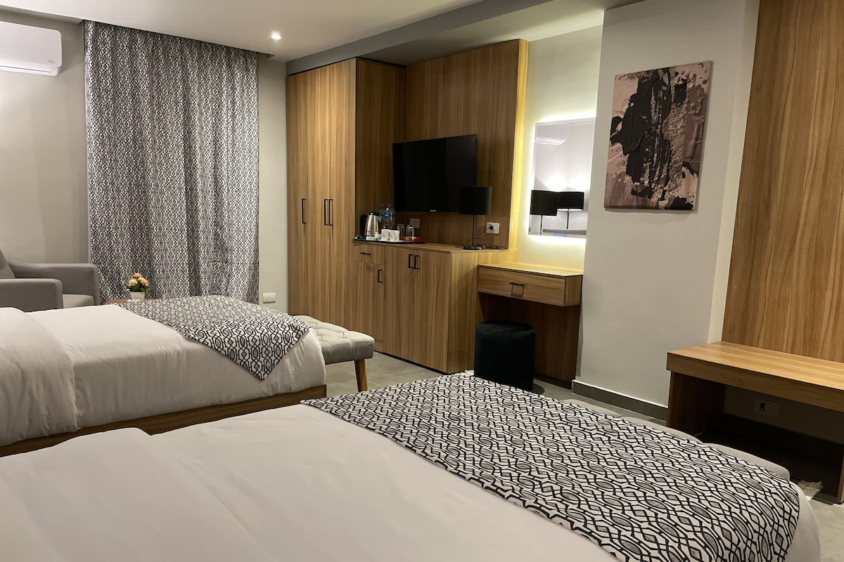 A hotel room with two beds, a desk, a TV, and a kitchenette.