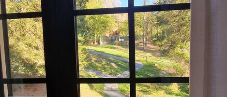 Vista a través de una ventana con una reja metálica, un balcón con baranda de metal y un sendero que conduce a una zona verde con árboles.