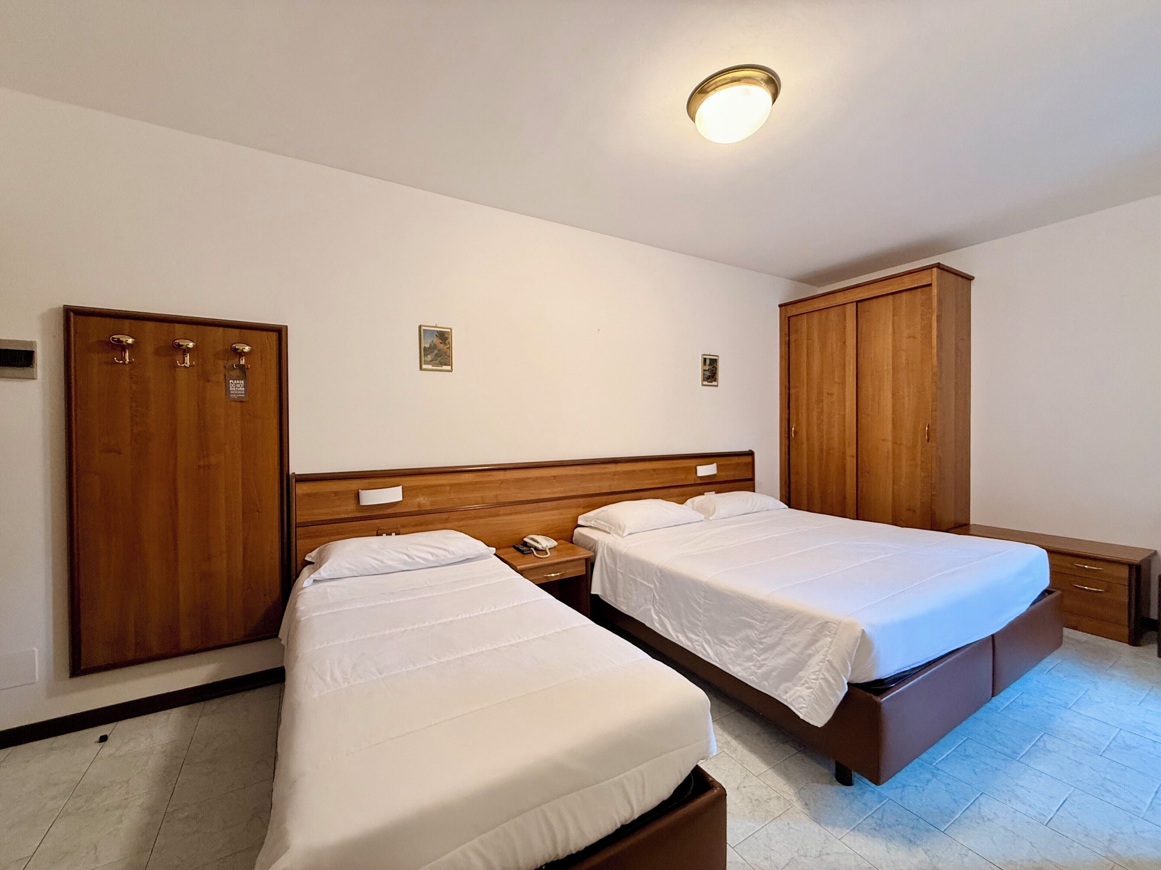 Habitación de hotel con dos camas individuales, armarios de madera, una mesita de noche con lámpara y un aplique de techo.
