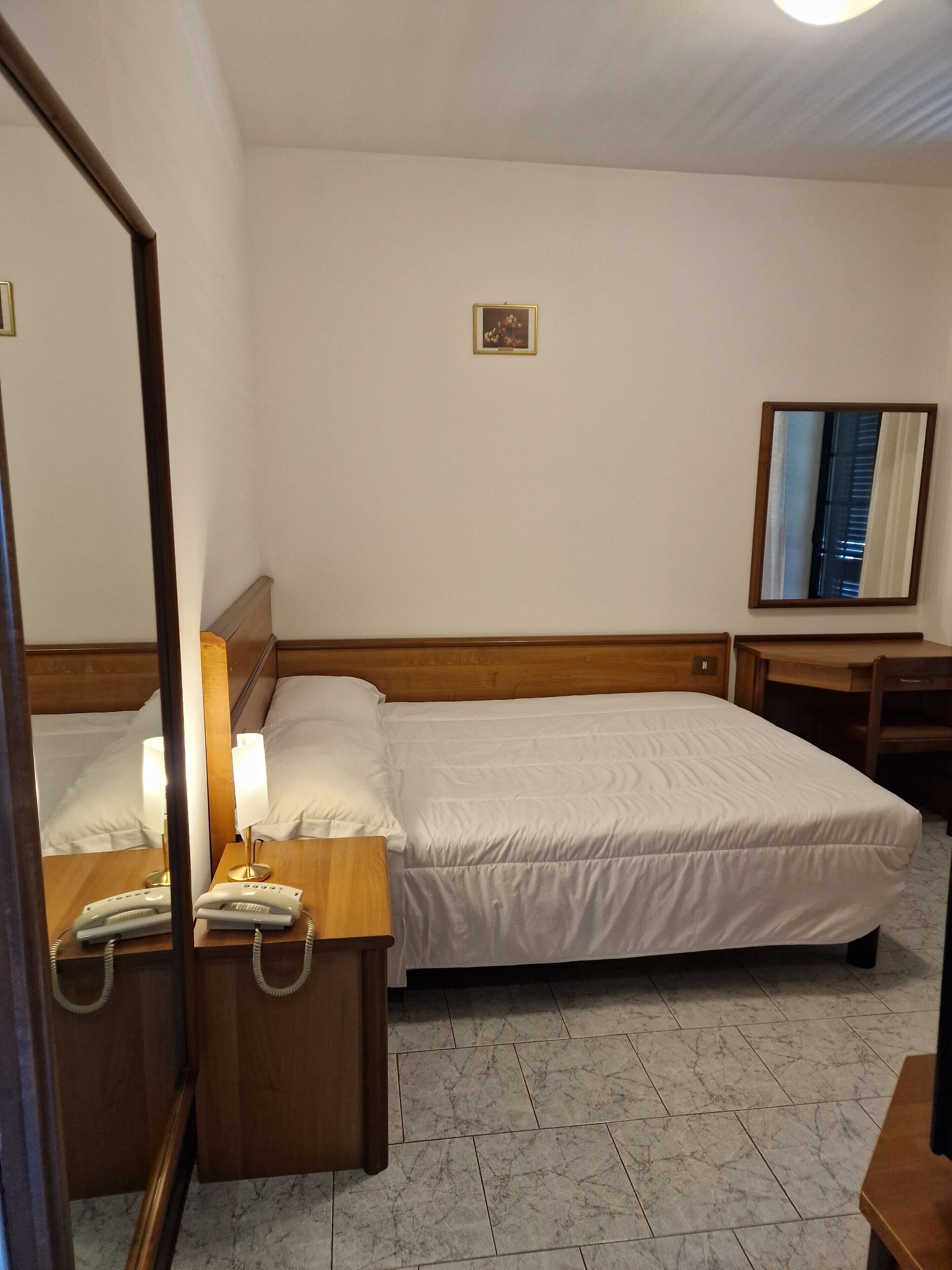 A hotel room with a bed, bedside tables, a mirror, a small picture on the wall, and a desk with a chair.