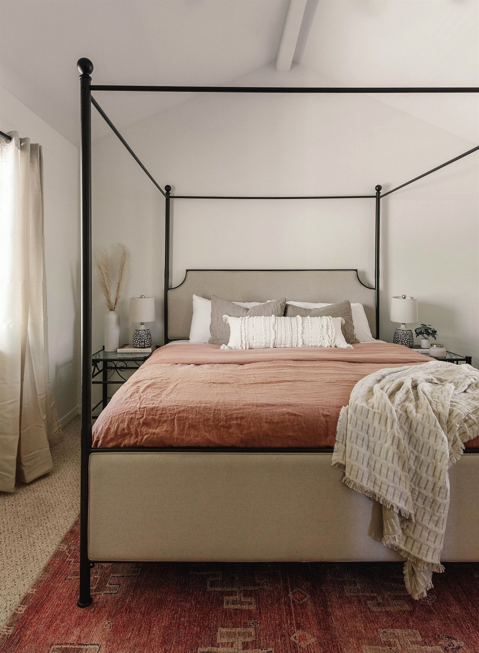 Un lit à baldaquin avec une tête de lit beige et une couvre-lit marron. Il y a deux tables de chevet avec des lampes et un vase sur l’une d’elles. La pièce est dotée d’un tapis à motifs et d’une fenêtre avec des rideaux.