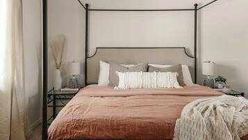 Un lit à baldaquin avec une tête de lit beige et une couvre-lit marron. Il y a deux tables de chevet avec des lampes et un vase sur l’une d’elles. La pièce est dotée d’un tapis à motifs et d’une fenêtre avec des rideaux.