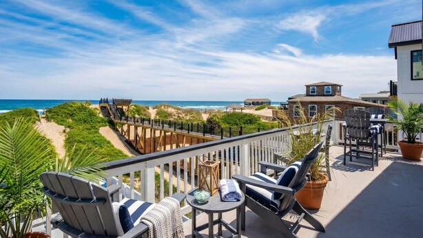 A balcony with outdoor seating, a view of the beach, and a wooden deck leading to the beach.