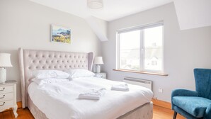 A bedroom with a large bed, a nightstand, a blue chair, a window with a view, and a painting on the wall. - Summercove 04 - Family Estate (Lahinch)