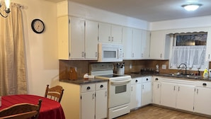Une cuisine avec des meubles blancs, un four à micro-ondes, une cuisinière et un évier. On y trouve une table à manger avec des chaises et une horloge au mur.