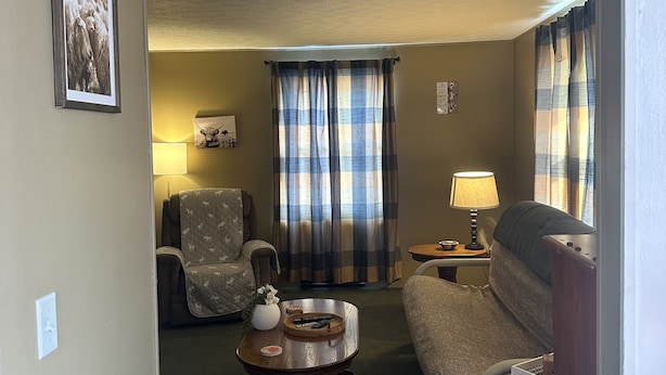 Un salon avec un canapé, un fauteuil et une petite table ronde sur laquelle se trouve une lampe. Il y a des rideaux et un tableau encadré au mur.