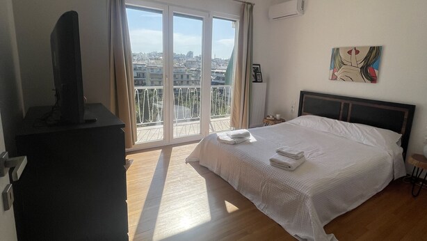 A bedroom with a large bed, a flat-screen TV, a ceiling fan, and a view of the city through large windows. - Pagrati Flat 7 (Αθήνα)
