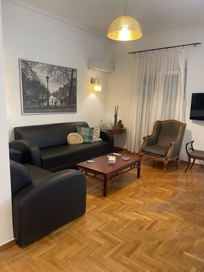 Un salon avec un canapé noir, une table basse en bois, un fauteuil à motifs et un téléviseur fixé au mur.